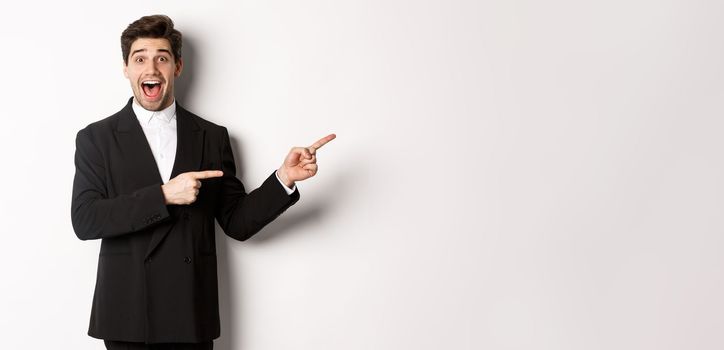 Image of attractive smiling guy dressed for new year party, pointing fingers right and showing advertisement, looking impressed standing over white background.