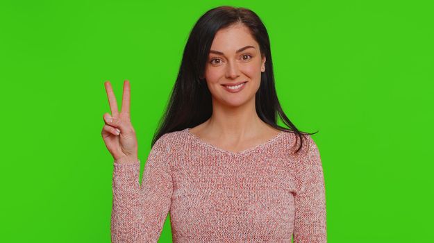 Hipster pretty woman in sweater showing victory v sign, hoping for success and win, doing peace gesture, smiling with kind optimistic expression. Young adult girl isolated on chroma key background