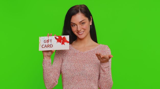 Young woman in sweater showing pointing on gift discount certificate voucher coupon for store holidays sale. Lovely girl with shopping christmas surprise card isolated alone on chroma key background