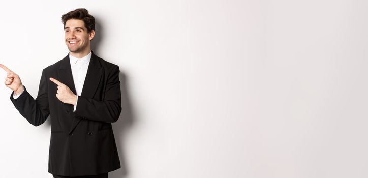 Portrait of successful handsome man in suit, pointing and looking left with pleased smile, showing promo banner, standing over white background.