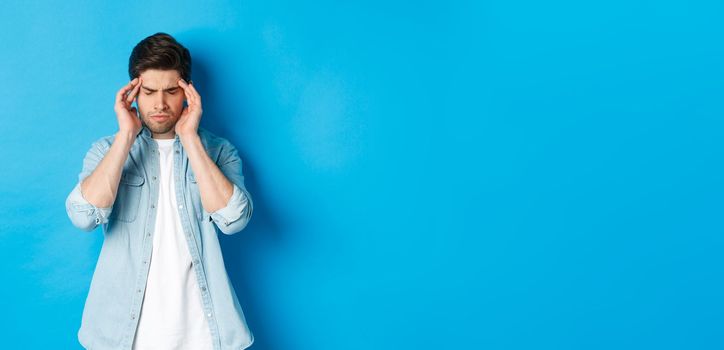 Man with hangover touching head and grimacing, having headache, standing against blue background.