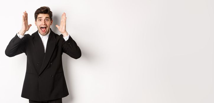 Frustrated and concerned man in black suit, screaming in panic and looking at something shocking, standing over white background.