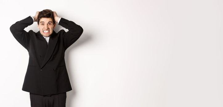 Image of frustrated and angry businessman in black suit, ripping hair on head and grimacing mad, standing tensed against white background.