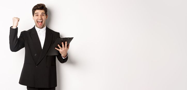 Portrait of excited handsome businessman in black suit, holding clipboard and making fist pump, achieve goal and rejoicing, standing over white background.