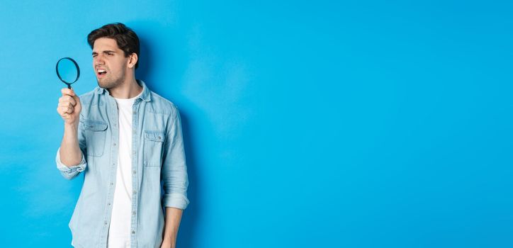 Young thoughtful guy looking through magnifying glass, reading something tiny, standing over blue background.