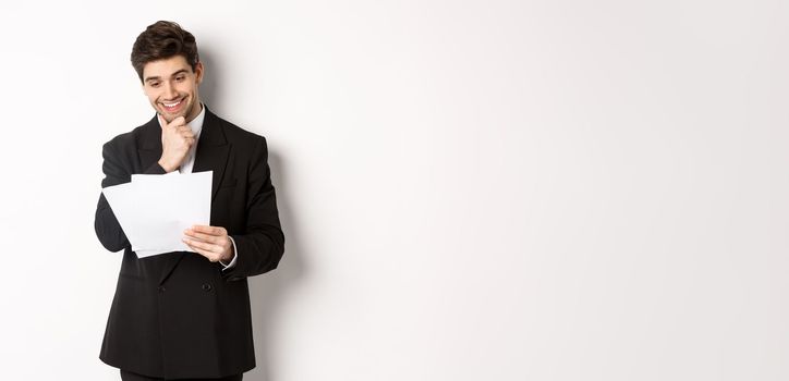 Image of handsome businessman in black suit, looking pleased at documents, reading report and smiling, standing against white background.