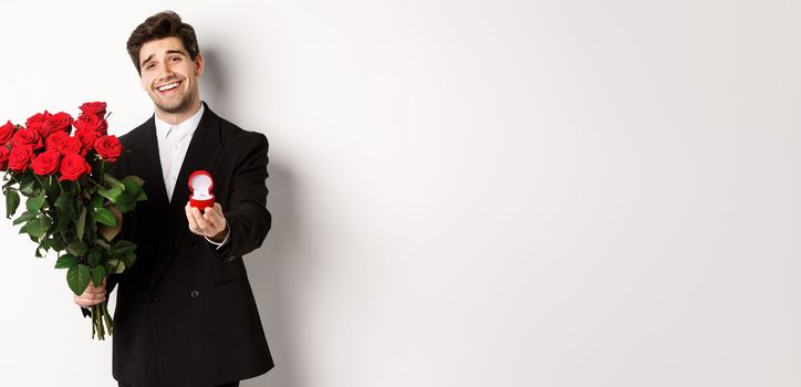 Handsome smiling man in black suit, holding roses and engagement ring, making a proposal to marry him, standing against white background.