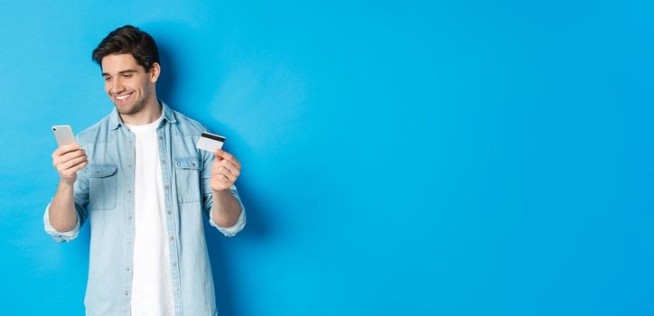 Young man shopping online with mobile application, holding smartphone and credit card, standing over blue background.