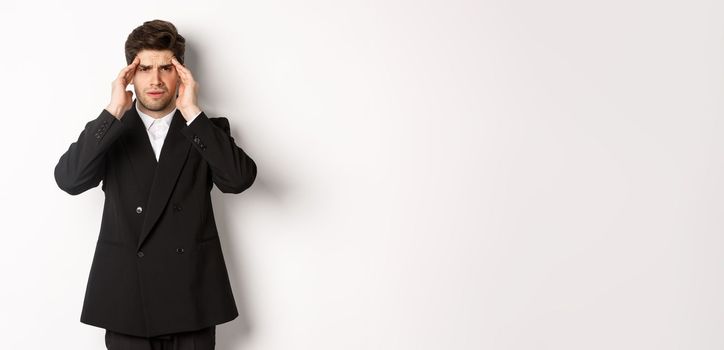 Image of businessman in black suit, touching head and looking dizzy, feeling painful headache, standing over white background.