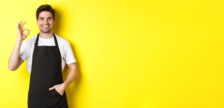 Young smiling barista in black apron showing okay sign, recommending coffee shop or restaurant, standing over yellow background.