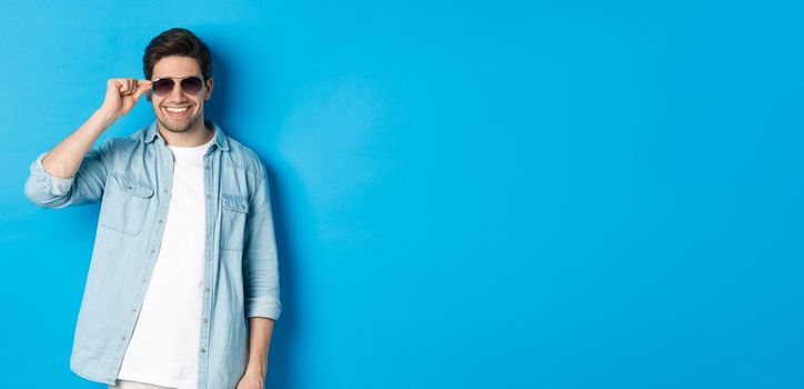 Confident macho man put on sunglasses, looking cool and sassy, standing over blue background.