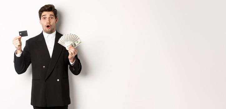 Portrait of surprised handsome man i suit, showing credit card with money, standing against white background.