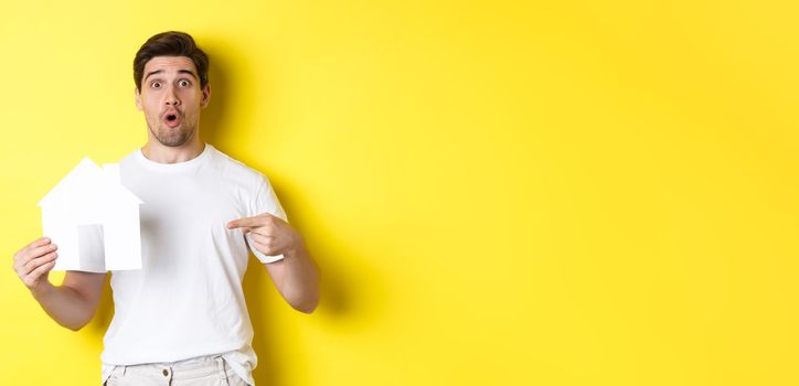 Real estate concept. Excited man pointing finger at paper house model, searching for flat, standing over yellow background.