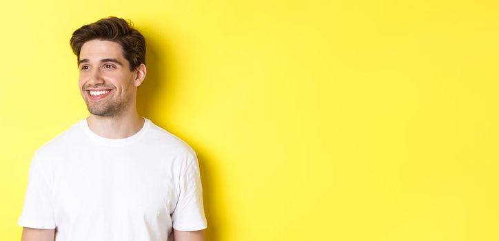 Close-up of attractive bearded man in white t-shirt smiling, looking left at copy space, standing against yellow background.