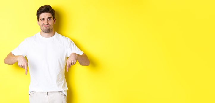 Young unhappy guy grimacing, pointing fingers down at advertisement, disappointed in product, standing over yellow background.