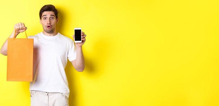 Surprised man showing mobile screen and shopping bag, standing against yellow background. Copy space