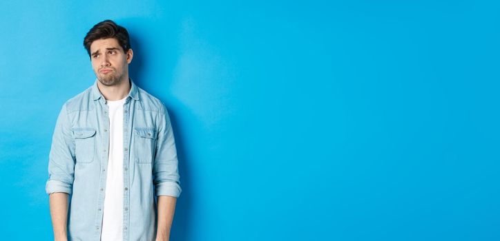 Image of reluctant and sad guy in casual outfit looking left, frowning and feeling upset, standing against blue background.