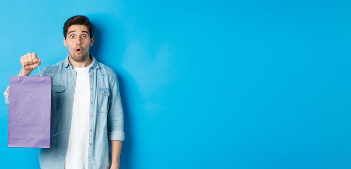 Concept of shopping, holidays and lifestyle. Handsome surprised guy holding paper bag from shop and looking amazed, standing over blue background.