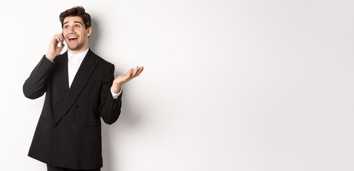 Portrait of happy good-looking businessman receiving great offer, talking on phone and looking pleased, standing in black suit against white background.