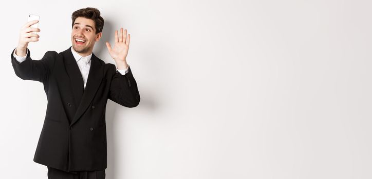 Image of handsome man in suit, having video call and waving hand at smartphone camera, recording video, greeting someone, standing against white background.