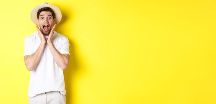 Concept of tourism and summer. Surprised male model in straw hat, looking amazed at special offer, standing against yellow background.