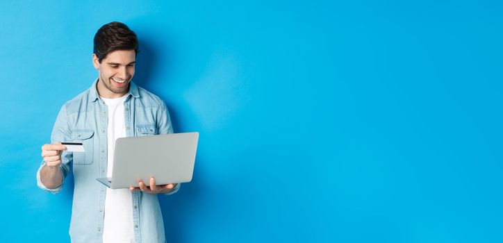 Young smiling man buying in internet, holding credit card and paying for purchase with laptop, standing over blue background.