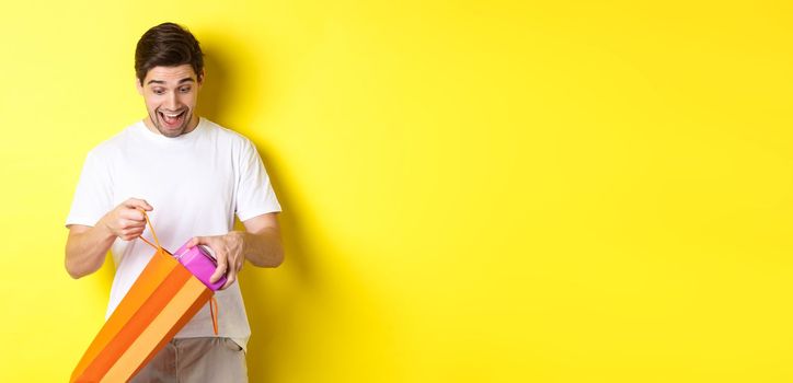 Concept of holidays and celebration. Young man looking surprised as take out gift from shopping bag, standing over yellow background.