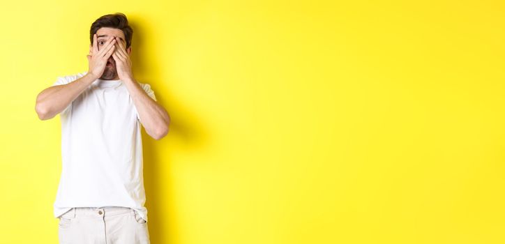 Embarrassed guy shut eyes but peeking through fingers at something awkward, standing over yellow background.