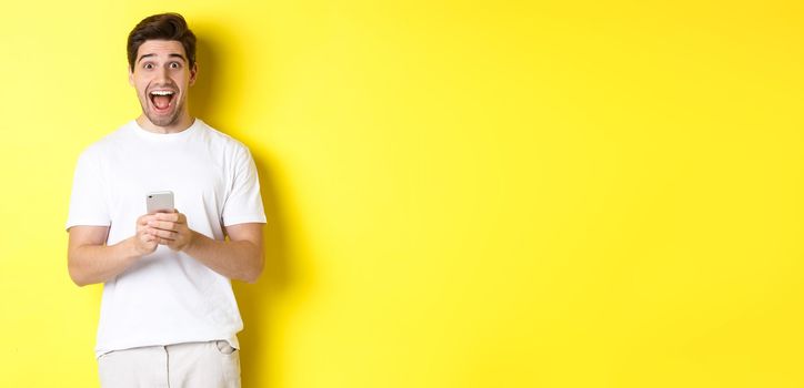 Man looking amazed and happy after reading something on mobile phone, standing over yellow background. Copy space