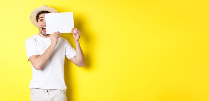 Excited tourist on vacation showing blank piece of paper for your logo, holding sign near face and smiling, standing against yellow background.