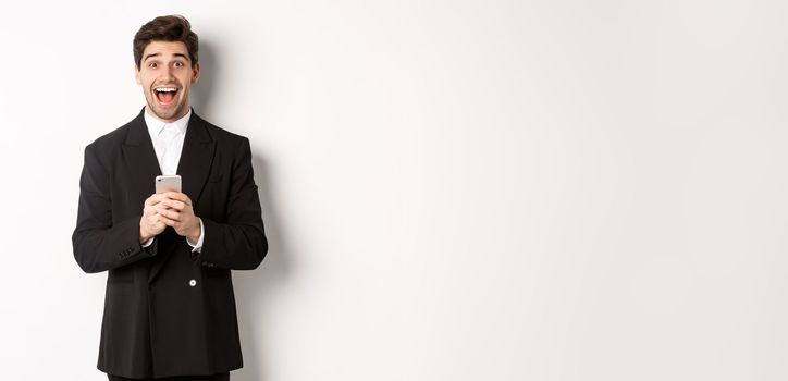 Image of excited handsome guy receiving promo offer on mobile phone, looking amazed and smiling, standing against white background.