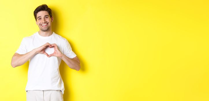 Happy romantic man showing heart sign, smiling and express love, standing over yellow background. Copy space