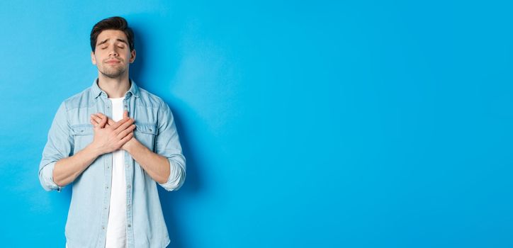 Portrait of dreamy and nostalgic guy in casual outfit, holding hands on heart and close eyes, daydreaming against blue background.