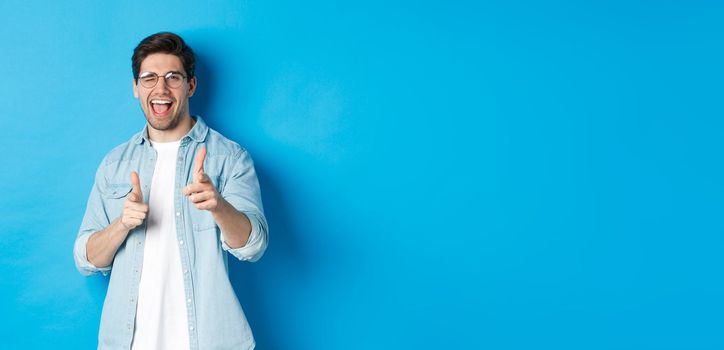 Confident man saying congrats, winking and pointing at you, standing pleased over blue background and smiling.