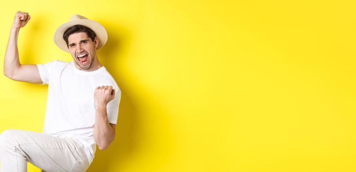 Concept of tourism and vacation. Lucky male tourist winning trip, rejoicing and saying yes, making fist pump while triumphing, standing against yellow background.