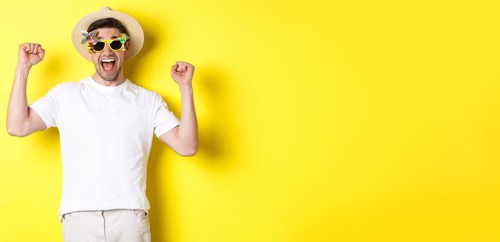 Concept of tourism and lifestyle. Happy man winning trip to resort, shouting yes and raising hands up, triumphing, wearing sunglasses and summer hat, yellow background.