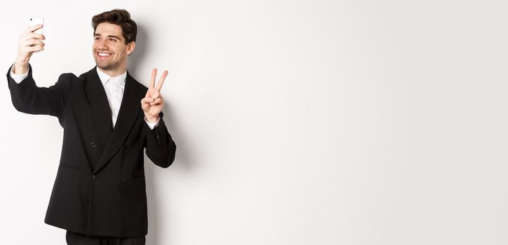 Portrait of good-looking man taking selfie on new year party, wearing suit, taking photo on smartphone and showing peace sign, standing against white background.