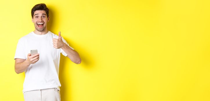 Happy smiling man holding smartphone, showing thumb up in approval, recommend something online, standing over yellow background.