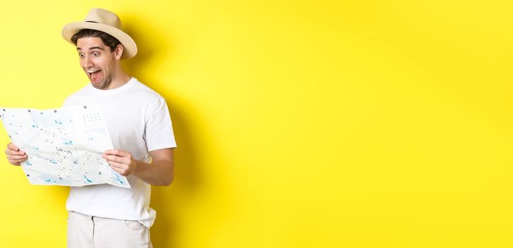 Travelling, vacation and tourism concept. Smiling happy tourist looking at map with sightseeings, standing against yellow background.