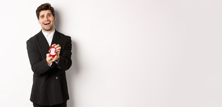 Portrait of handsome man in black suit, open box with wedding ring, making a proposal, asking to marry him, standing against white background.