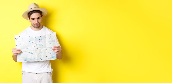 Travelling, vacation and tourism concept. Male tourist looking confused at map, looking for right direction, standing over yellow background.