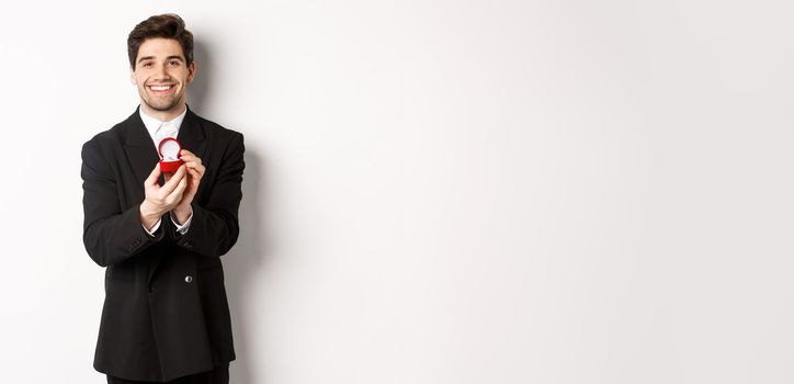 Image of handsome boyfriend in black suit making a proposal, asking to marry him and showing wedding ring, standing over white background.