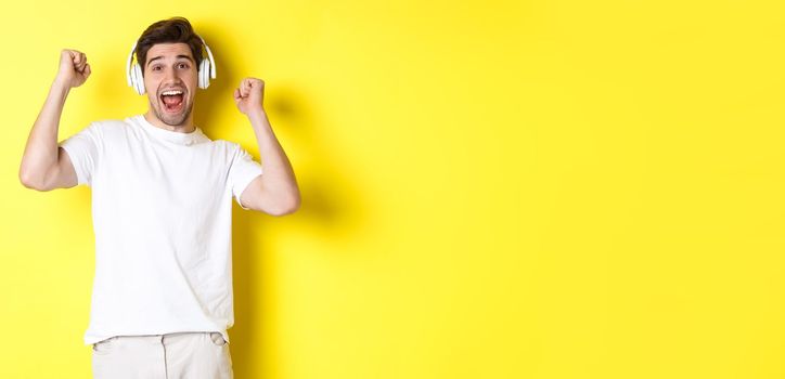 Excited handsome man dancing and singing along, listening music in headphones, standing over yellow background.