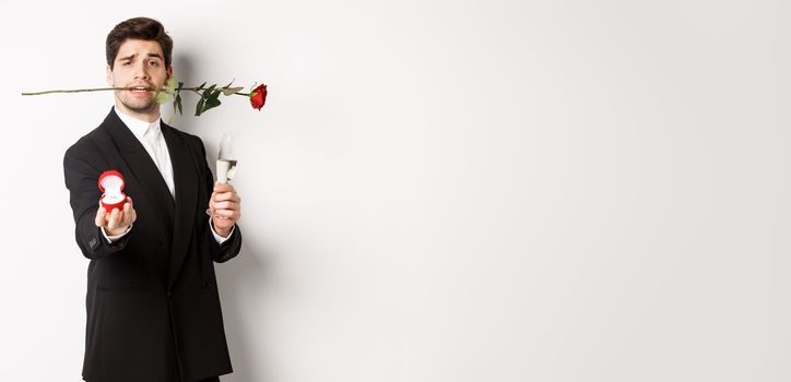 Romantic young man in suit making a proposal, holding rose in teeth and glass of champagne, showing engagement ring, asking to marry him, standing against white background.