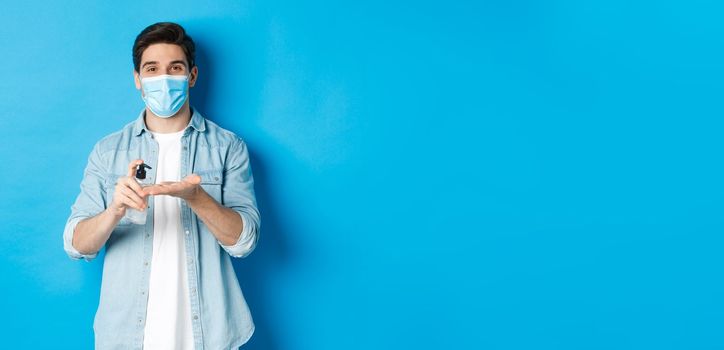 Concept of covid-19, pandemic and social distancing. Handsome young man in medical mask disinfecting hands with sanitizer, using antiseptic, standing against blue background.