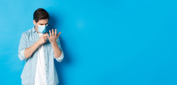 Concept of coronavirus, social distancing and pandemic. Man in medical mask looking at his palm through magnifying glass, standing over blue background.