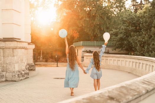 Daughter mother run holding hands. In blue dresses with flowing long hair, they hold balloons in their hands against the backdrop of a sunset and a white building