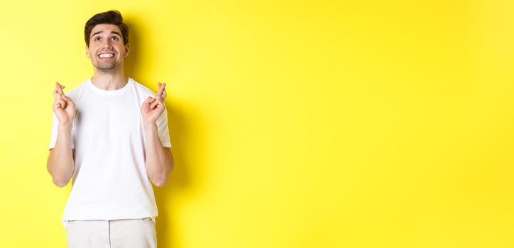 Nervous and hopeful man praying to god, making wish with fingers crossed, panicking and standing over yellow background.