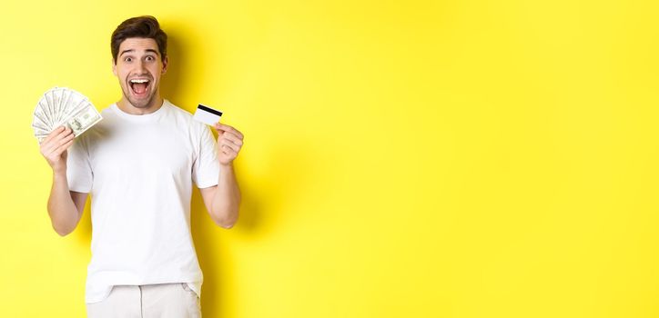 Excited man ready for black friday shopping, holding money and credit card, standing over yellow background.