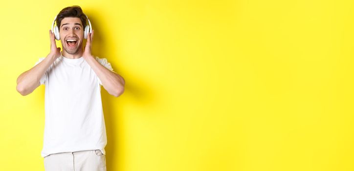 Man in headphones looking surprised and happy, listening awesome song, standing over yellow background.